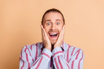 Excited young man holding his face in surprise against a beige background, showcasing an expressive...