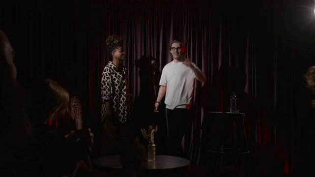 African American woman asking her colleague to come up on dimly lit stage for his stand-up performance