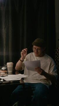 Vertical shot of smiling man sitting at table and rereading and making notes on paper in his stand-up performance script