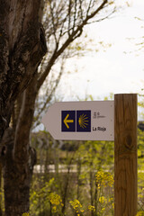 Wayfinding sign on the Camino de Santiago with a yellow arrow and scallop shell, pointing left toward La Rioja. A symbol of pilgrimage and direction in a spring forest setting.