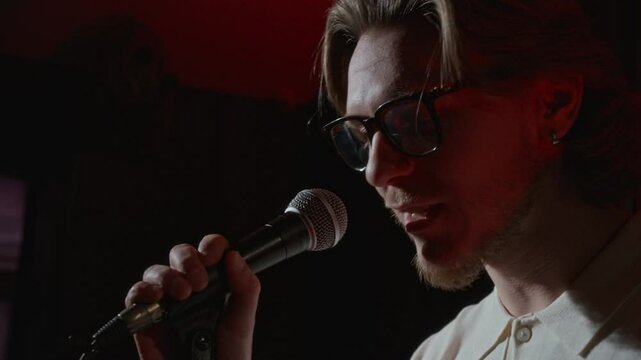 Close-up shot of young man in glasses talking in microphone on dimly lit stage with fading red light