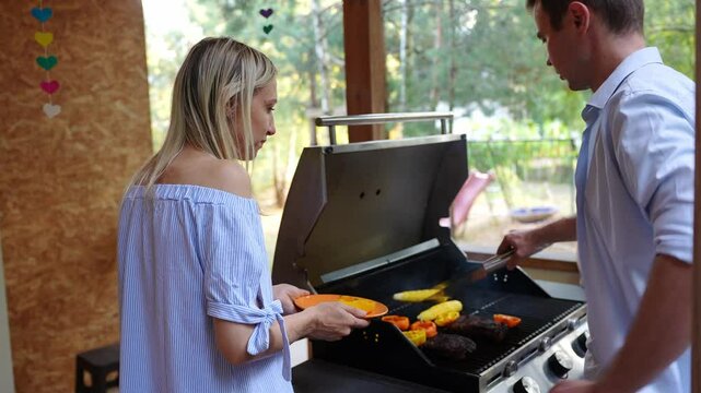 Man , woman cooking delicious food together on grill on veranda of house outdoors, girl watches as mom takes vegetables off grill. Family enjoys communication, cooking together. Family barbecue party.