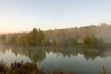 Morning fog on the lake at dawn. Colourful glow in the sky, with surrounding forest trees reflected in the calm water, in an atmospheric landscape.