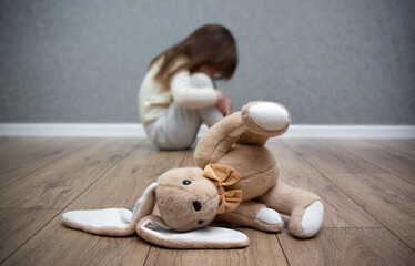 A stuffed bunny on the floor, a scared child sitting behind it. Child abuse. Bullying in the family.