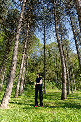 Obraz premium A girl in a black top and pants, with red shoes, stands in a forest, looking up at the tall trees surrounding her. The sun filters through the leaves.