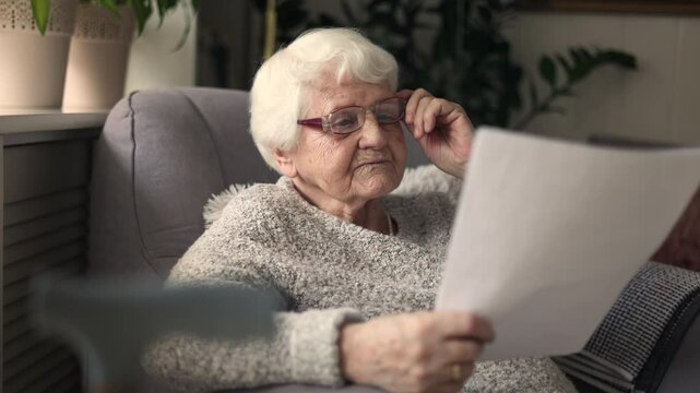 Elderly woman wearing eyeglasses reading documents while sitting in an armchair in living room