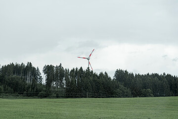 Fototapeta premium Wind turbine spins gracefully in a cloudy landscape surrounded by lush green trees