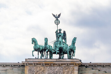 The Brandenburg Gate in Berlin features a majestic statue of a chariot led by four horses, symbolizing peace and victory against a cloudy sky backdrop.