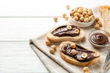 Slices of bread with chocolate spread and hazelnuts on white wooden table, closeup. Space for text