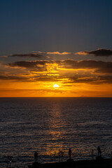 The sun is setting over the ocean, casting a warm glow on the water. A few people are standing on the beach, enjoying the beautiful view. The sky is filled with clouds