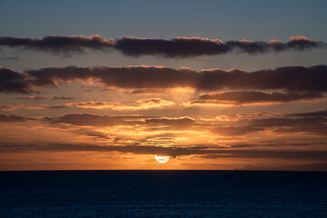 The sun is setting over the ocean, casting a warm glow over the water. The sky is filled with clouds, creating a serene and peaceful atmosphere
