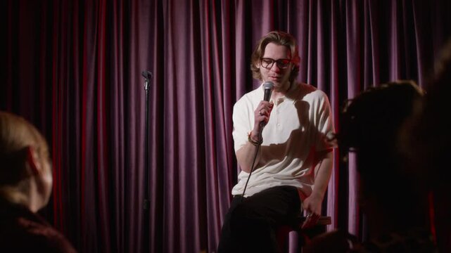 Young man in glasses giving stand-up performance on stage, talking in microphone, while audience clapping