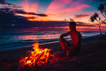 Man sitting by a beach bonfire at sunset, enjoying peaceful solitude and stunning ocean view.