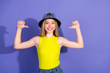Fototapeta premium Young woman in a yellow top and hat making gestures on purple background conveys a cheerful and stylish lifestyle