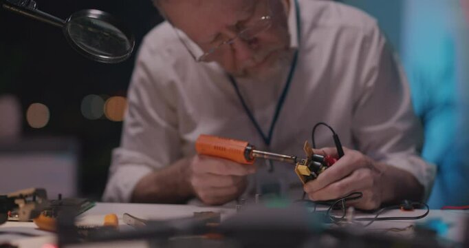 Adult engineer working late in office repairing microship motherboard with aoldering iron modern equipment. MAnufacturing. Mechanical repair.