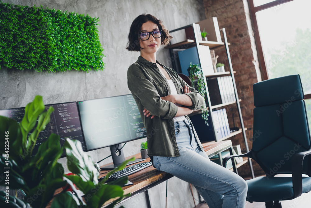 Poster confident female programmer standing next to her workspace decorated with greenery and coding monito