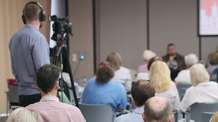 A videographer recording a formal conference presentation with an engaged audience.