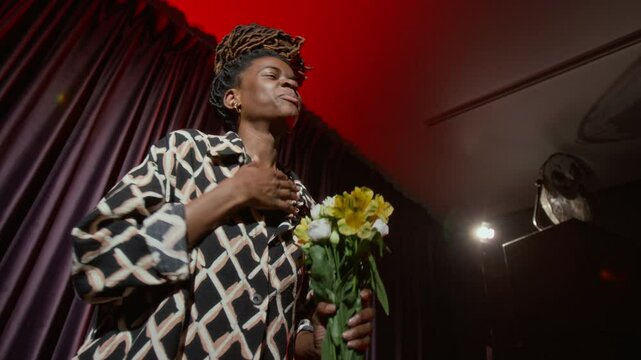 Low angle shot of African American woman with flowers bowing on dimly lit stage, thanking audience