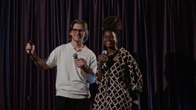 Slow motion shot of male and female stand-up artists smiling while standing on stage and holding microphones