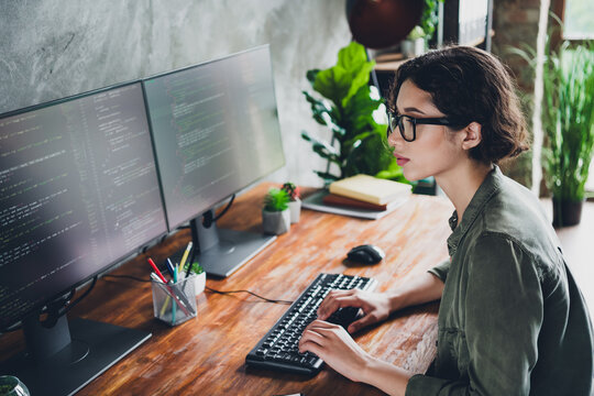 Young female programmer working at her desk with dual monitors, coding computer software in a well lit home office