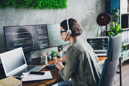 Young female programmer working at office desk with multiple screens, wearing casual attire, focused on coding and programming