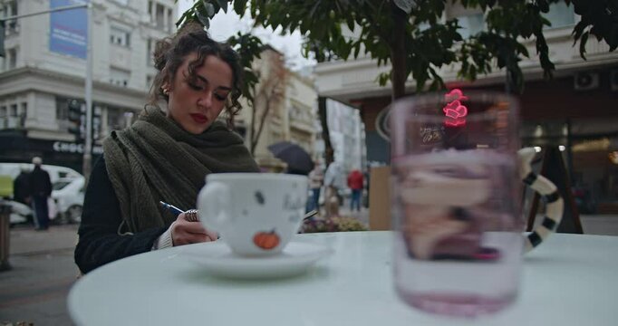 A young woman sits at a cafe table in the city, writing in her notebook and enjoying a cup of coffee.