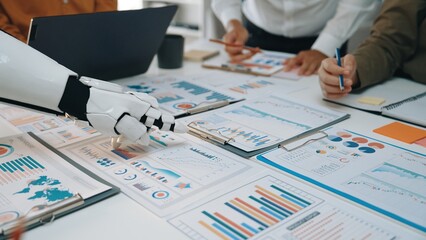 A modern business scene featuring a robotic hand assisting a professional team in analyzing charts, graphs, and reports on a collaborative workspace.Noogenesis
