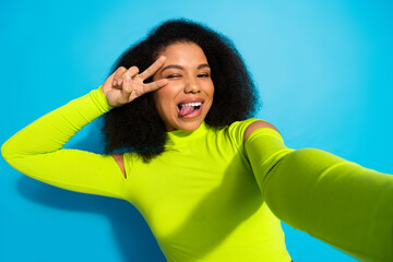 Vibrant young woman making peace gesture with cheerful expression on bright blue background showcasing neon green fashion outfit