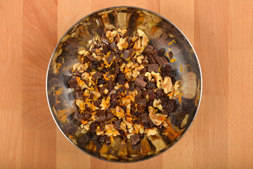 A top-down view shows a stainless steel bowl containing a mixture of walnuts, chocolate chunks, raisins, and grated orange and lemon zest on a light brown wooden surface
