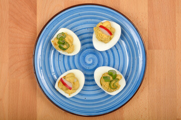 A top-down view shows four deviled eggs, filled with a creamy avocado and yolk mixture and garnished with green onions and radish slices, arranged on a blue swirled plate on a wooden surface