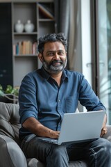 Young thoughtful bearded smart indian professional business man executive looking away sitting on chair in modern office lobby using laptop, thinking of new ideas, planning project, working online.