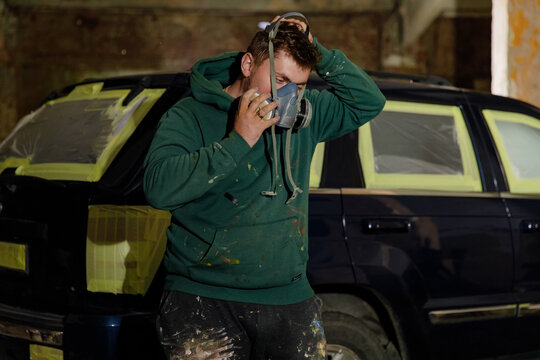 A man is engaged in car restoration, adjusting his gas mask while wearing a paint-splattered hoodie in a dimly lit workshop. Various tools and fixtures fill the space around him. - Powered by Adobe
