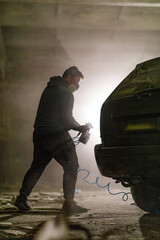 In a dimly lit workshop, a man wearing a mask is intently spray painting the rear of a car. Dust...