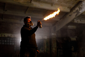 An artist wearing a gas mask ignites a spray can to create a mesmerizing flame, showcasing their unique skills in an abandoned warehouse setting at night.
