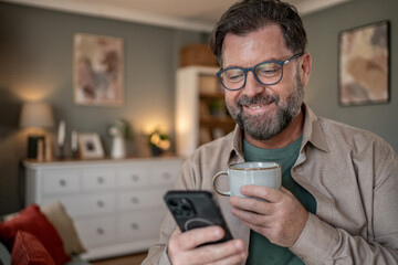 Smiling man using smartphone and drinking coffee at home