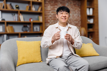 A man sits comfortably on a soft gray sofa with bright yellow cushions, joyfully sipping tea in an...