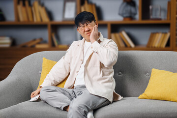 severe toothache, Upset man, holds his cheek, man sits cross-legged on a gray sofa, visibly...