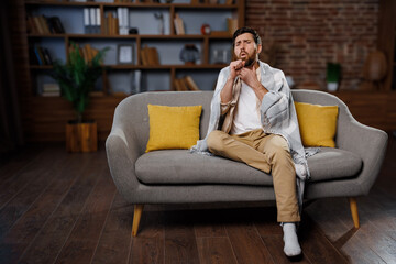 man coughs, feeling unwell, cold throat, man sits comfortably on a gray sofa, wrapped in a soft...