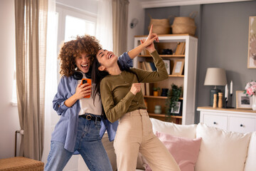 Cheerful Young Women Dancing Together In Living Room While Using Smartphone And Having Fun At Home