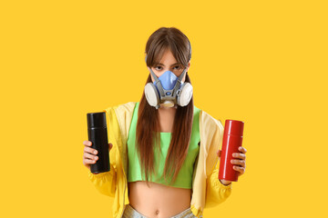 Young girl in respirator and with spray cans on yellow background