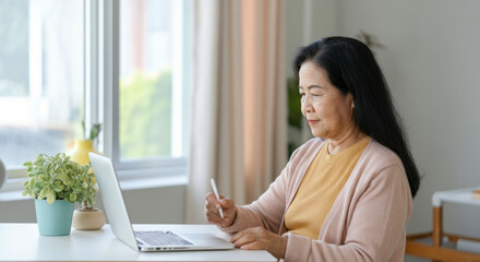 Elderly asian female using laptop and stylus in bright living room setting