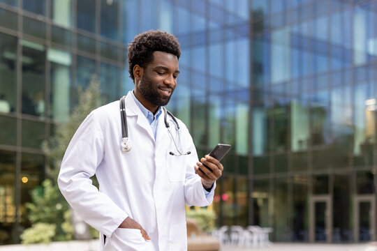 Successful doctor walking outside modern clinic office with phone in hand. Hospital worker using online app on smartphone, smiling contentedly. - Powered by Adobe