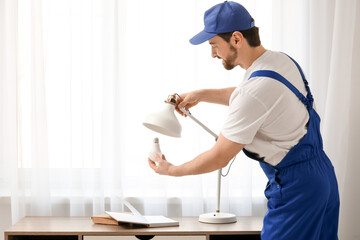 Happy male electrician changing light bulb in lamp on table near window at home