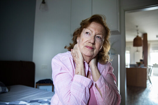 Senior woman with neck pain sitting on bed in home bedroom