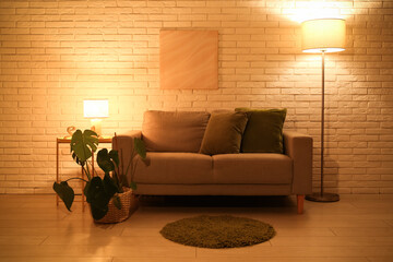 Interior of night living room with sofa, glowing lamps and houseplant on stool near white brick wall