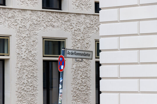 Street sign for An der Kommandantur positioned on a pole beside a contemporary building with unique textured walls in an urban setting.
