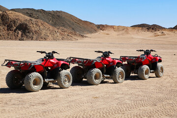Red quad bikes lined up on desert terrain with sand dunes and rocky hills in the background