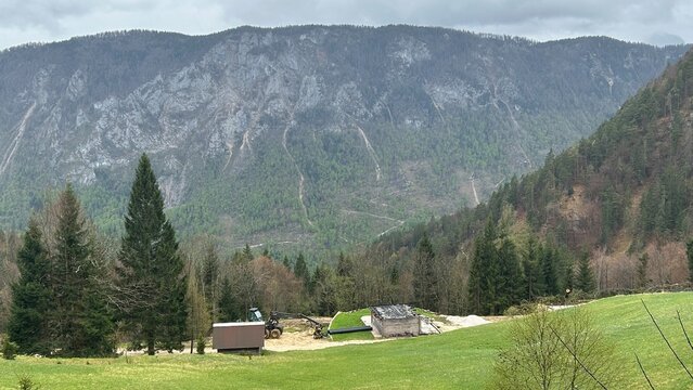Alpine scene of green meadow hill and high rock packs at the background with rural cabins and elements of construction in the center