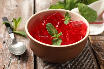 Delicious red jelly cubes in bowl, mint and spoon on wooden table, closeup