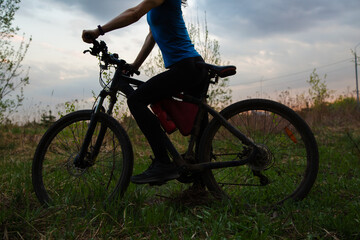 Fototapeta premium Beautiful sport body woman with strong legs riding on bicycle on the green grass and looking on the nature sunset spring background. Sporty wear female girl outdoors